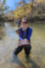 A young woman in waders fly fishing in a stream showing off a rainbow trout
