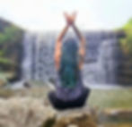 Person with long hair sits cross-legged on rocks, arms raised, meditating in front of a waterfall. Peaceful nature setting.