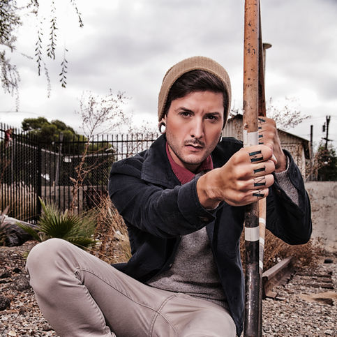 Low-angle editorial portrait of a man with a beanie and a denim jacket, crouching and holding an arrow with hand tattoos, set against an industrial fence. This Chiaroscuro shot emphasizes movement and composition.