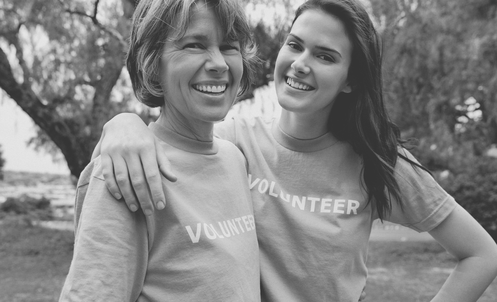 Older Caucasian woman smiling with a younger Caucasian woman, both wearing volunteer shirts, photo is black-and-white