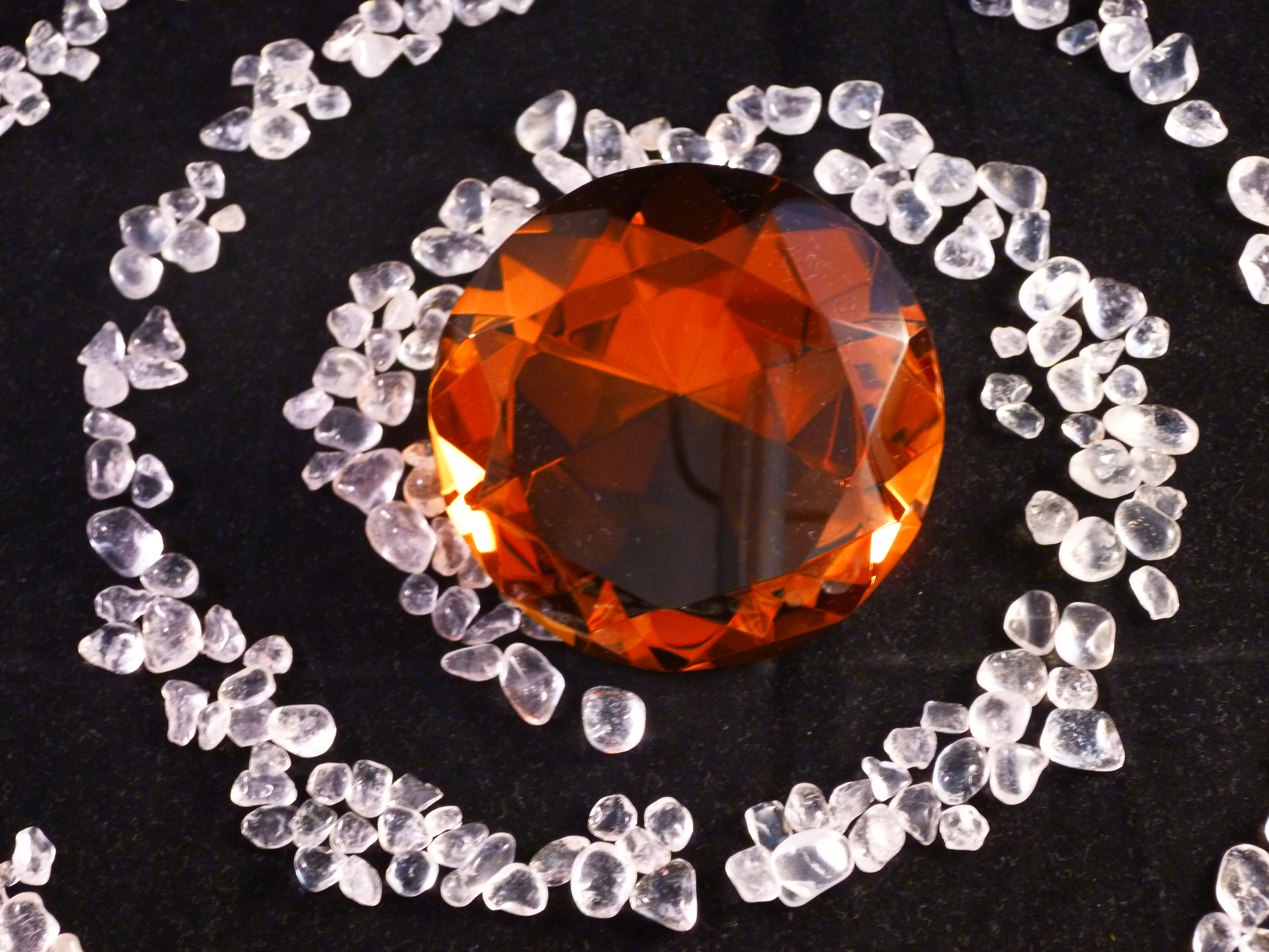 Orange Kristallstein auf schwarzem Hintergrund, umgeben von kleinen Steinen. Amaris.