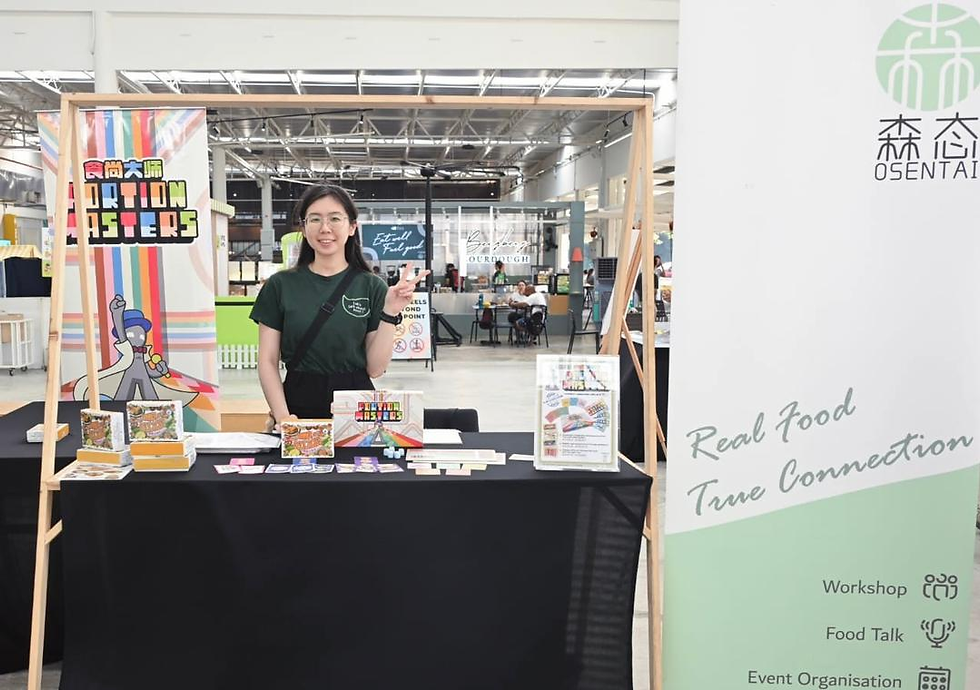 展台上,一名穿绿色T恤的女性微笑,比出胜利手势。旁边有“Portion Masters”海报,背景为明亮的市场环境。