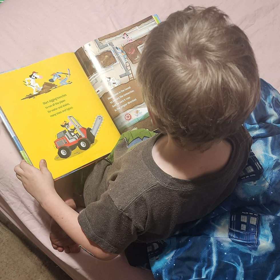 Young child sitting on a bed with blue blanket, reading a colorful book. The book shows illustrations of dogs and construction vehicles. Calm mood.