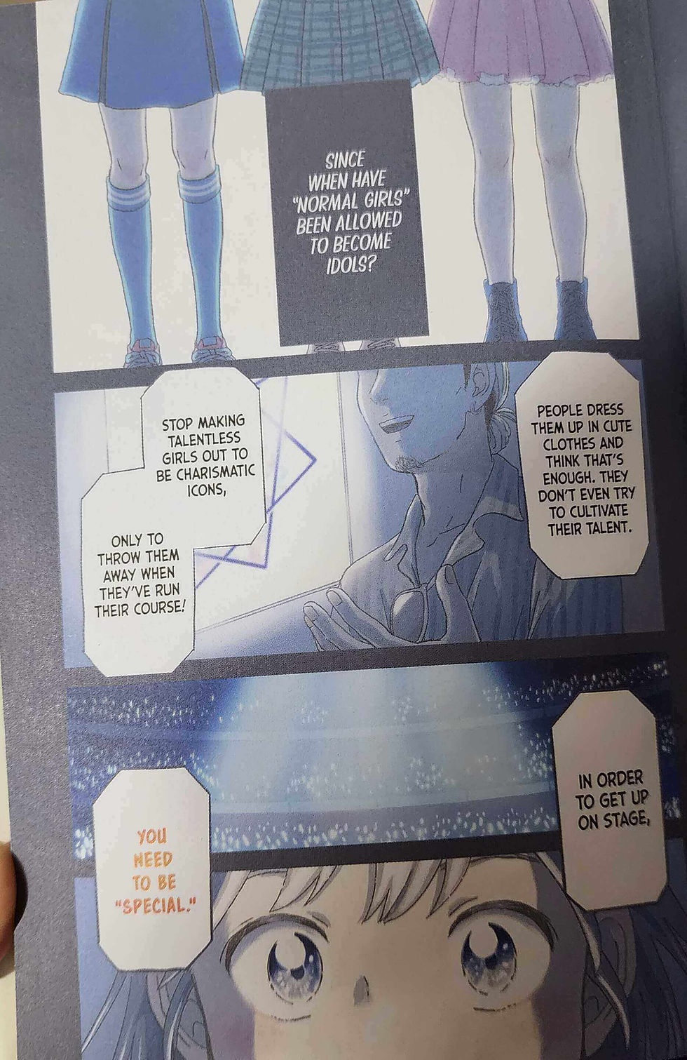 Manga panel with girls' legs onstage and a smiling man; speech bubbles criticize idol culture, with spotlights and blue-purple tones.