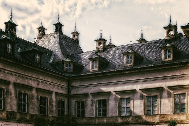Historic manor with steep slate roof, dormer windows, and spires under cloudy sky, giving a moody, weathered look.
