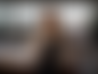 A woman in a blazer standing confidently in an office with colleagues working behind her.