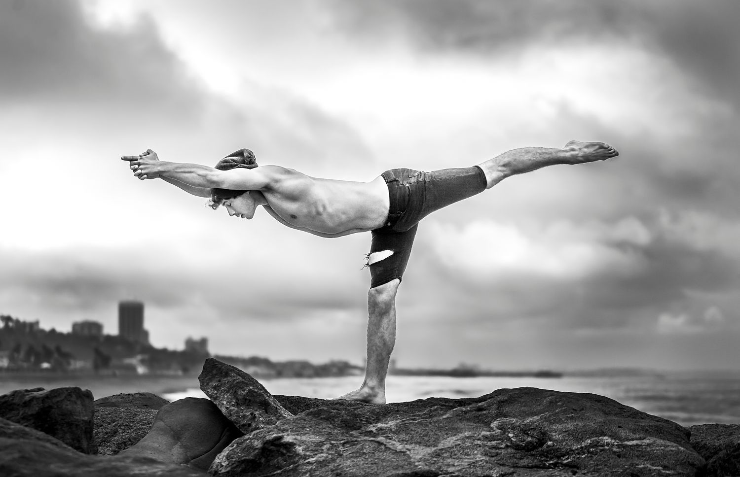 Black and white photo of dancer Jake Tedrick balancing on one leg on coastal rocks with arms extended forward and leg back, c