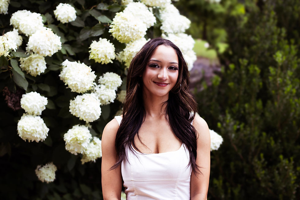 A young woman standing in front of a flower bush in a white dress.