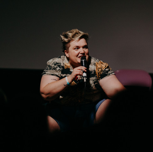 Lucy, a small round person with goblin energy, crouching on stage