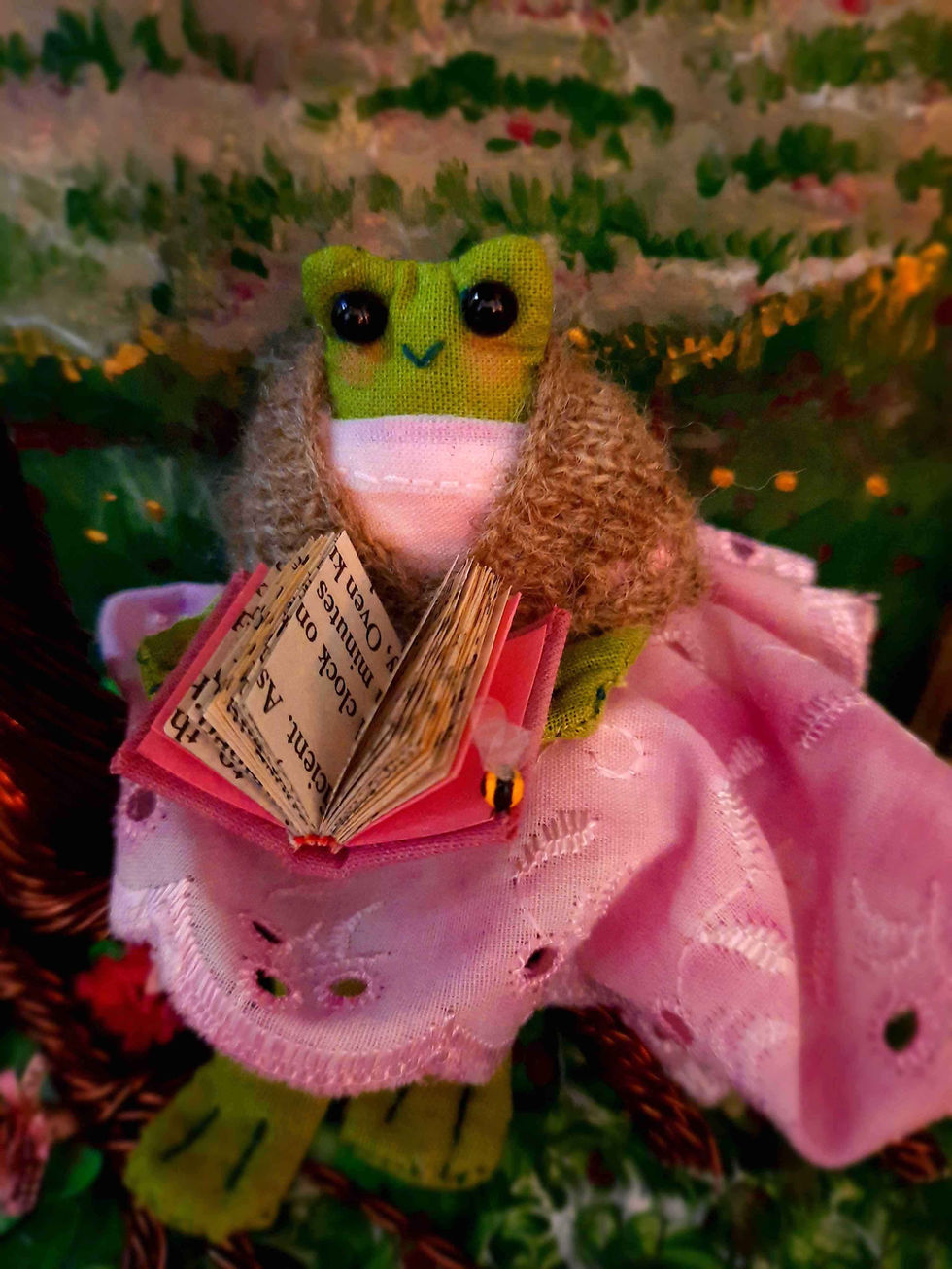 A stylised frog with black beaded eyes sitting on a tree root. The frog is wearing a pink dress and a knitted shawl, and holds an open book in her hands. A bee sits at the corner of the book.