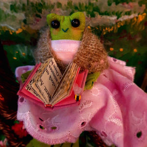 A stylised frog with black beaded eyes sitting on a tree root. The frog is wearing a pink dress and a knitted shawl, and holds an open book in her hands. A bee sits at the corner of the book.