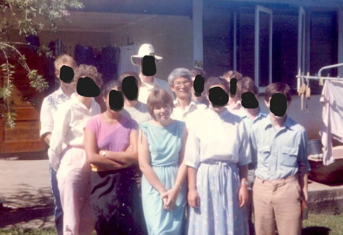 A group of adult men and women in the backyard of a home. 10 have their faces blacked out. An Asian man in his fifties with grey hair, glasses and a white collared shirt stands behind the 19-year-old writer, Wendy, who has short brown hair and has her hands clasped in front of her. She is wearing a blue dress