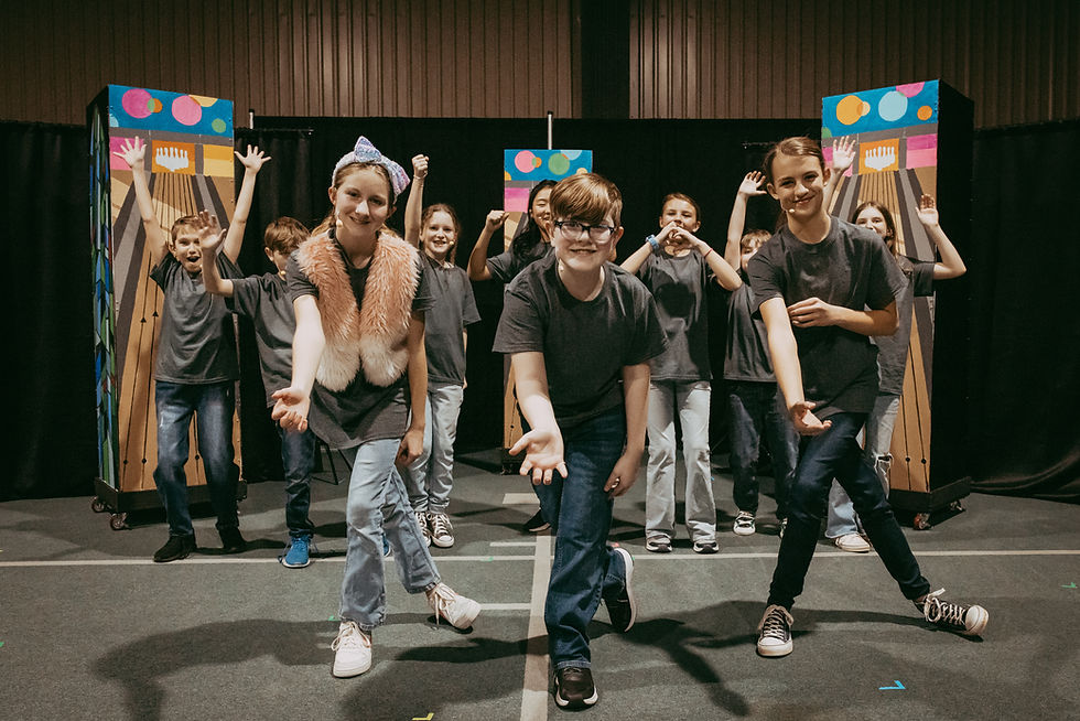 A group of children pose energetically on stage, wearing dark shirts and jeans. Bright backdrop panels add color. Happy mood.