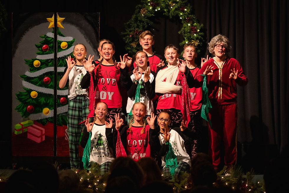 Kids in Christmas pajamas sing onstage, making OK hand signs, before a lit wreath and painted tree with gifts; festive, cheerful scene