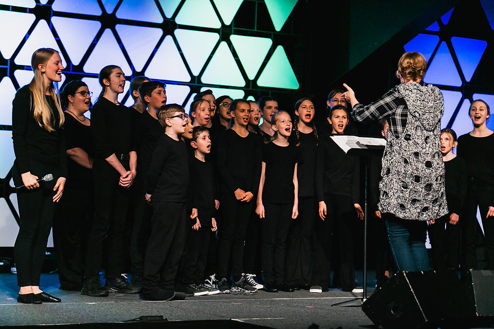 Choir of children in black singing on stage, guided by a conductor. Background features a geometric pattern with blue and green lights.
