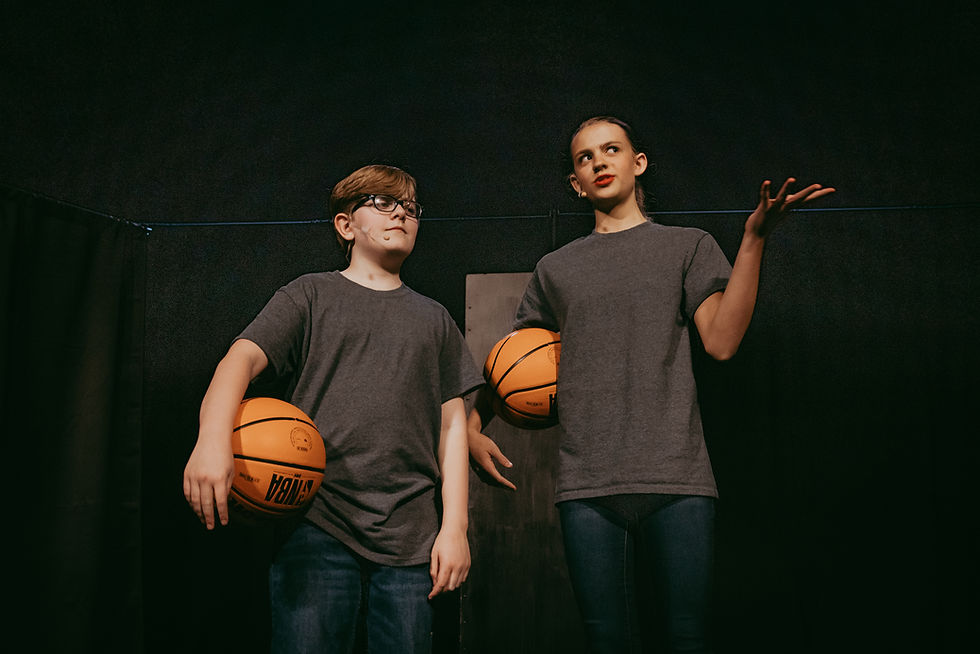 Two children in gray shirts hold basketballs on a dark stage. One gestures expressively. Mood is serious, but funny. 