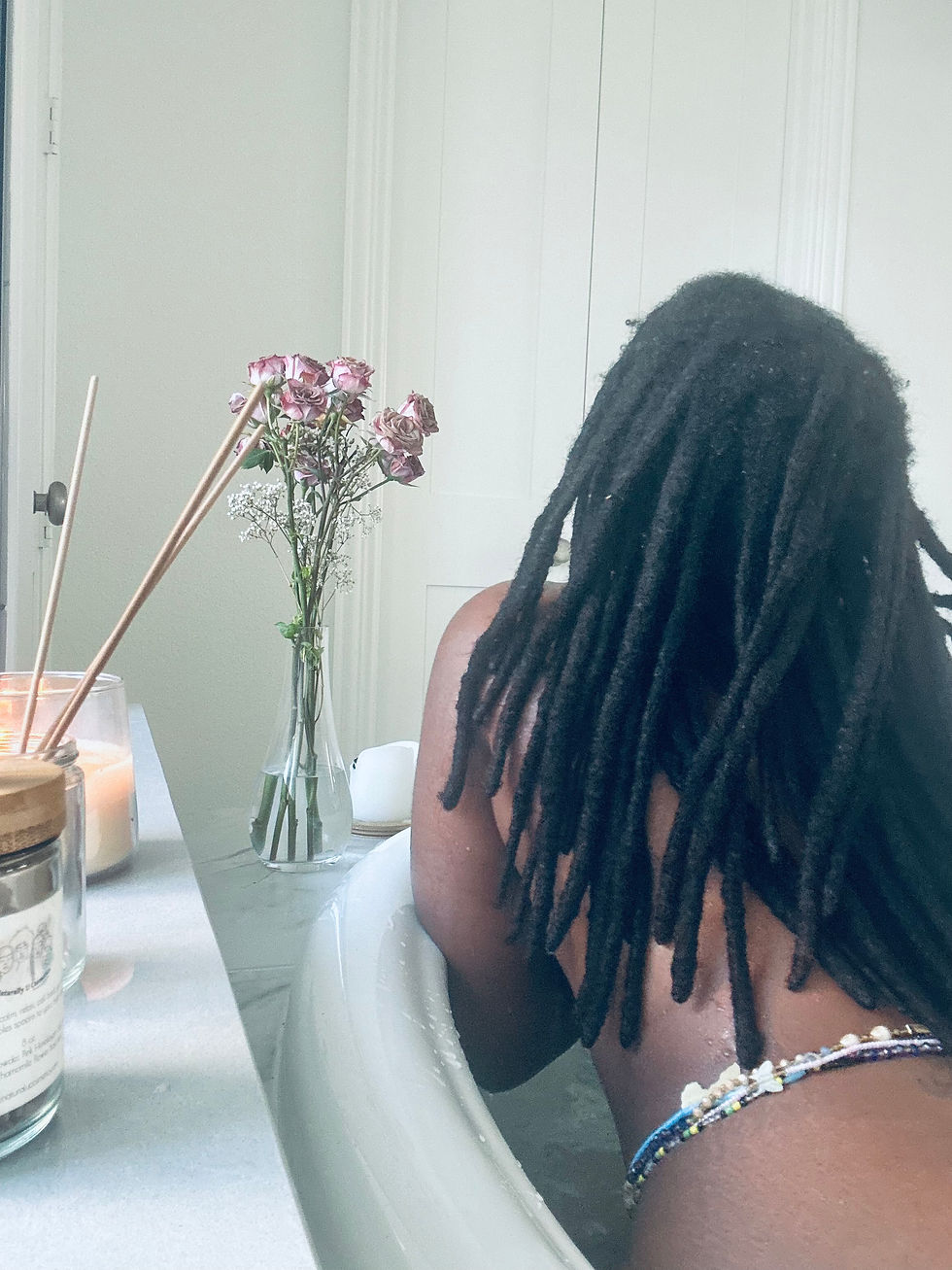 a black woman who face you can't see has locs and waist beads inside of a tub with a candle and roses.