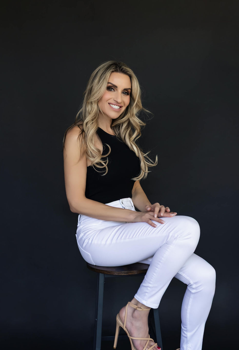 Smiling woman with long blonde hair seated on a stool. She wears a black top, white pants, and beige heels against a dark background.