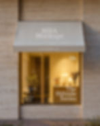 Exterior storefront mockup with limestone walls and a fabric awning, showing a warm interior with a lamp and flowers.
