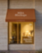Exterior storefront mockup with limestone walls and a fabric awning, showing a warm interior with a lamp and flowers.