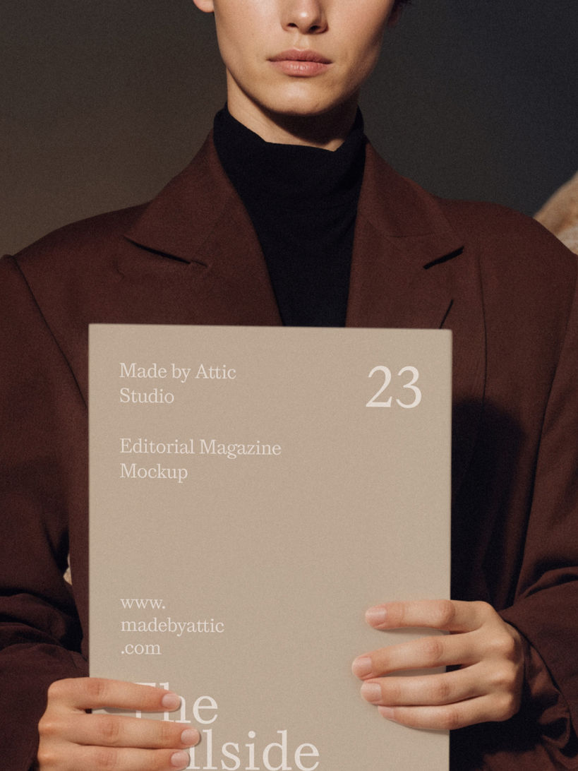 Model with short hair in a brown coat holding a blank white magazine cover next to a large textured rock.