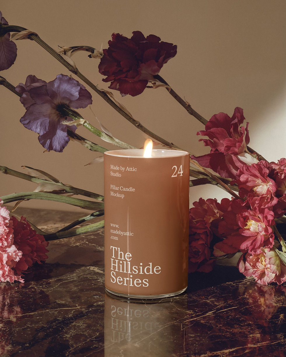 A high-fidelity pillar candle mockup with a glowing flame, set on a dark marble surface with purple and red flowers in the ba