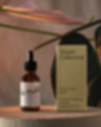 Amber glass dropper bottle standing next to a white packaging box on a stone pedestal with a pink leaf overhead.