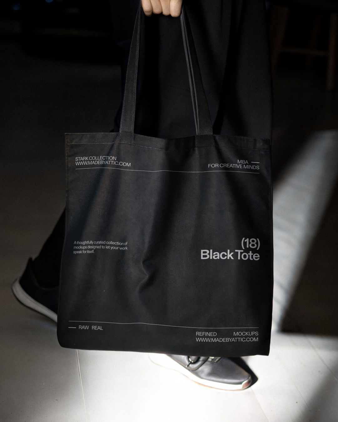 A high-fidelity black tote bag mockup held by a person in a moody, high-contrast urban setting.