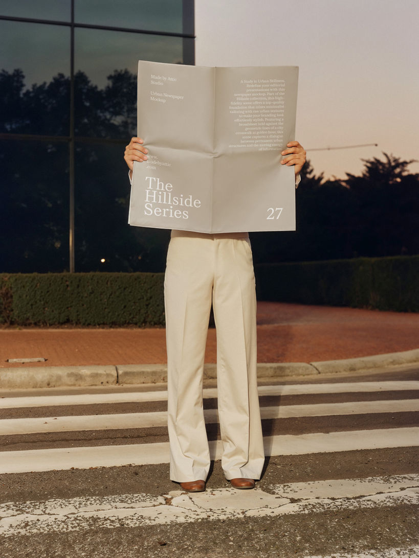 A high-fidelity newspaper mockup with a blank broadsheet page held by a person in cream trousers on an urban crosswalk during