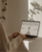 Close up of hands in a beige knit sweater typing on a laptop computer with soft leaf shadows cast on the white wall behind.