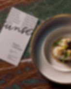 Vertical blank menu card lying on dark green marble next to a gourmet dish on a gold-rimmed plate.