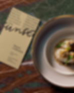 Vertical blank menu card lying on dark green marble next to a gourmet dish on a gold-rimmed plate.