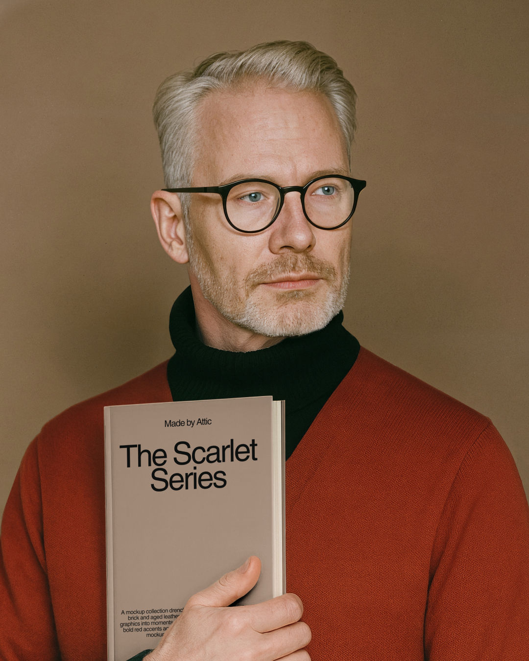A high fidelity book mockup held by a distinguished man with grey hair and glasses wearing a red cardigan over a black turtle