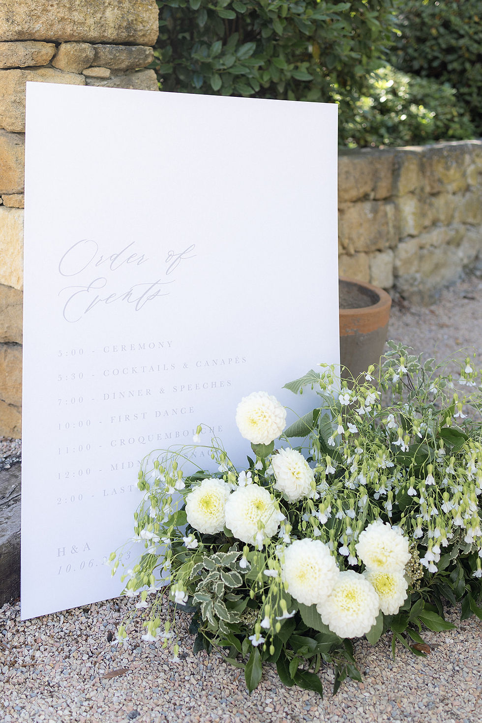 An order of events sign designed with calligraphy on white card, styled next to a white and green foliage floral arrangement.