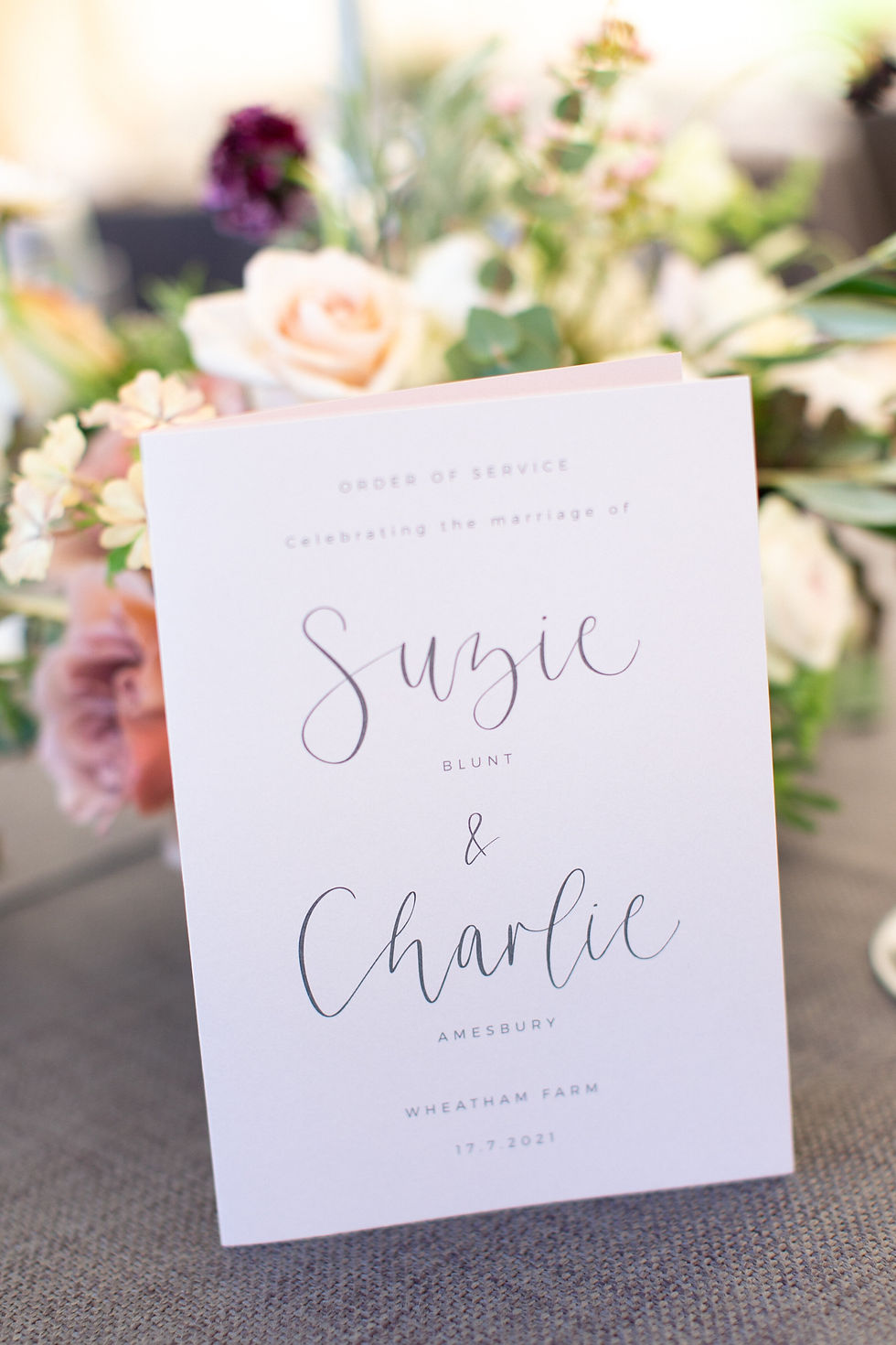 Wedding order of service booklet with modern calligraphy on white card, positioned in front of beautiful pastel summer flowers