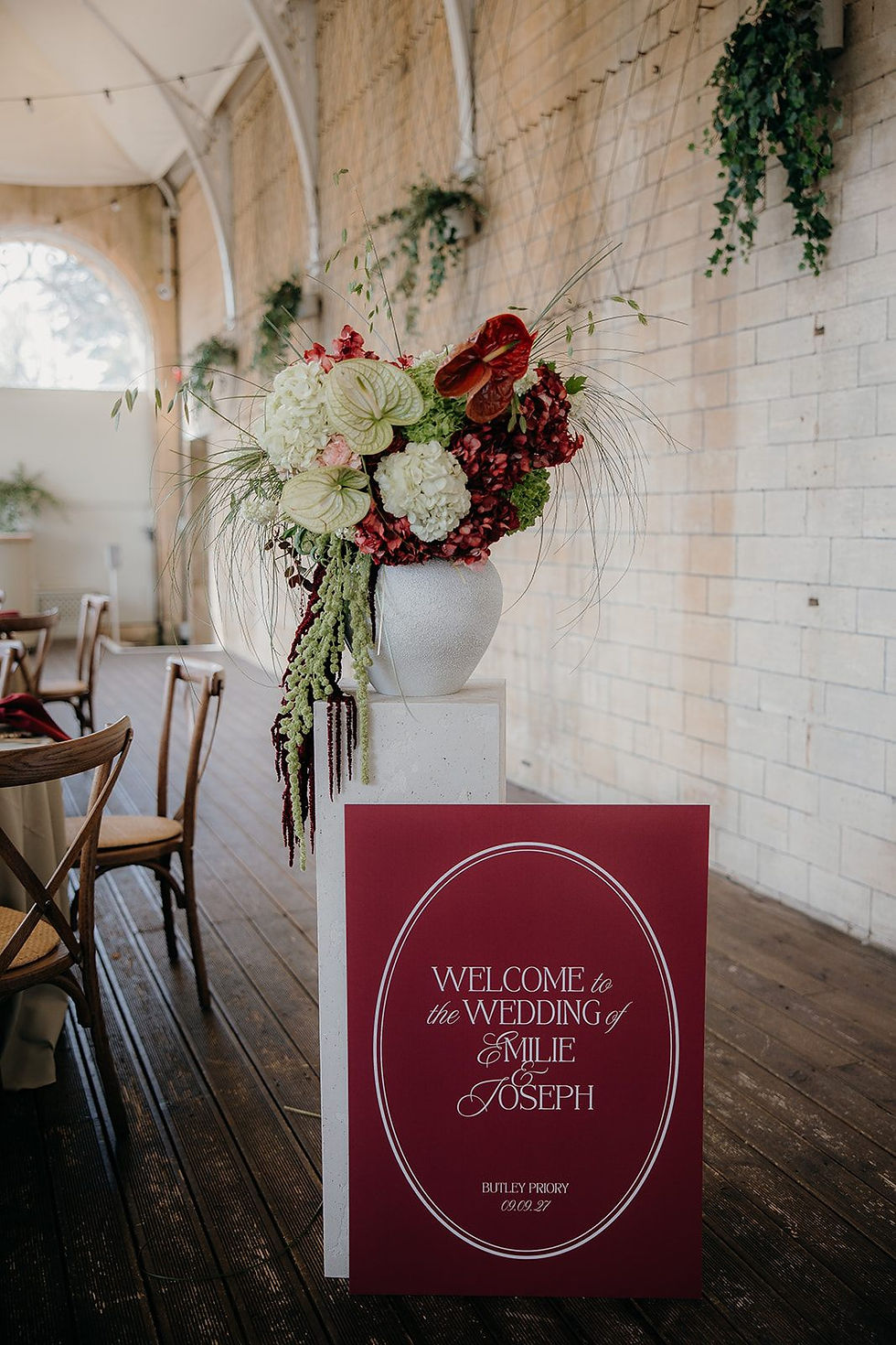 A wedding welcome sign on burgundy card with white text, styled next to stunning wedding florals in green, cream and burgundy in a vase 