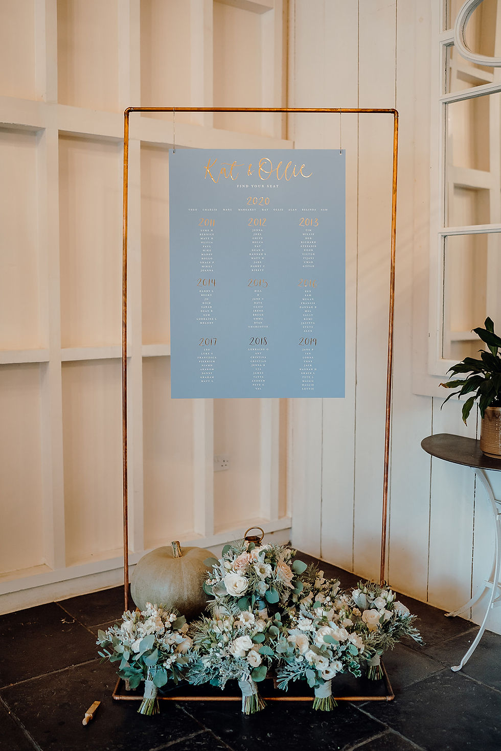 Wedding table plan on a pale blue background with white text and gold foil calligraphy, hung on a copper frame.