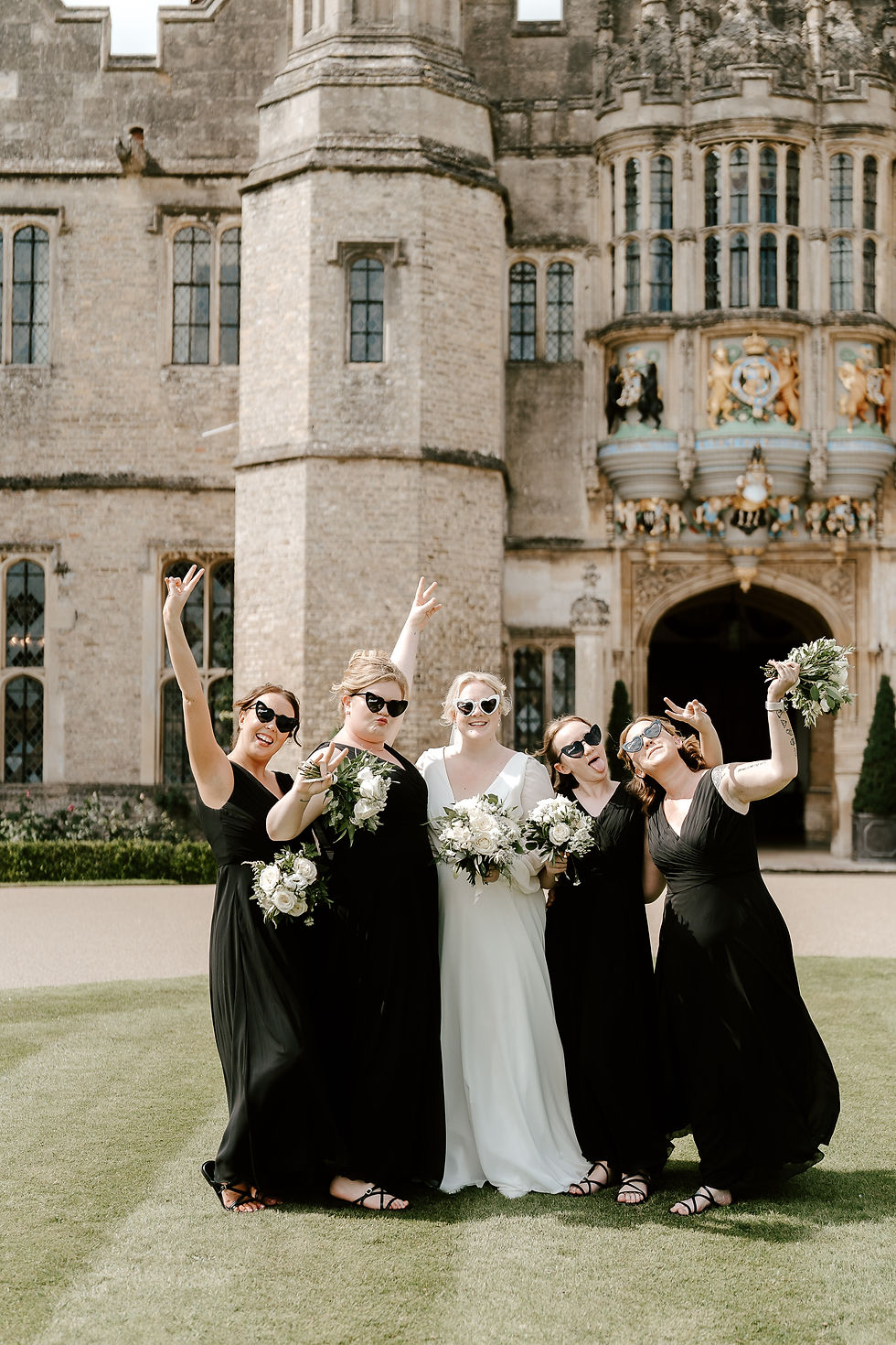 A bride and her four bridesmaids wearing black maxi dresses, pose outside a Tudor house wedding venue, all wearing heart sunglasses