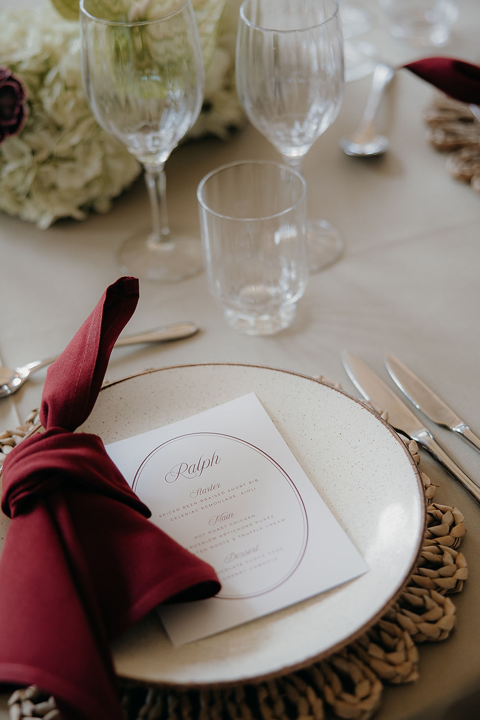 A wedding guest place name combined with a wedding meno on white card in an oval shape, placed next to a burgundy napkin.