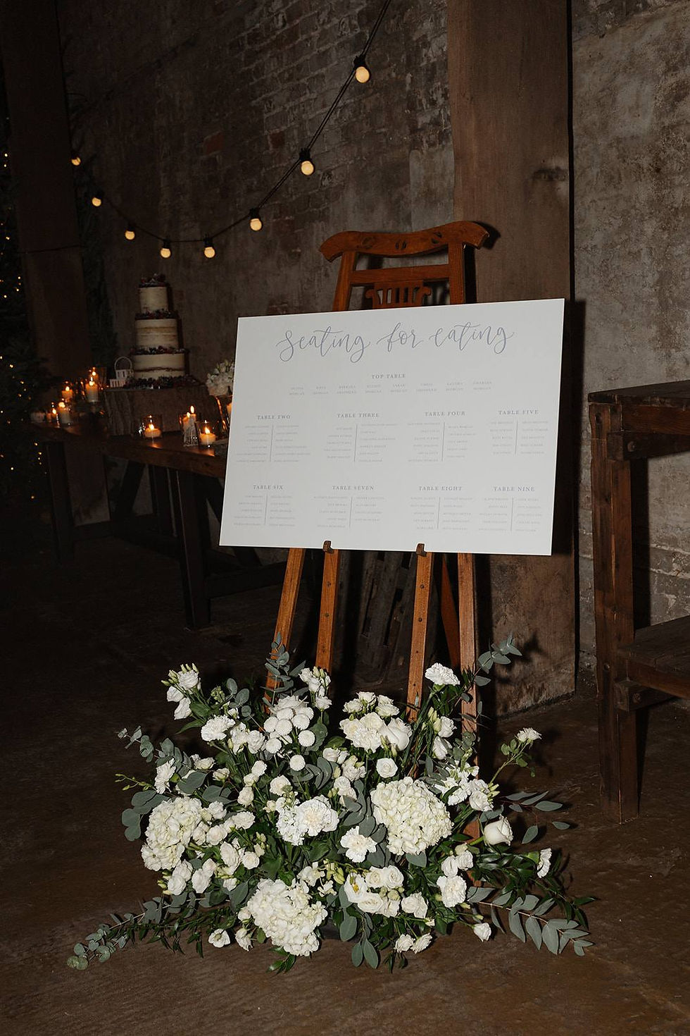A wedding table plan with a grey calligraphy header and grey text on a white background, placed on a wooden easel and styled with white flowers