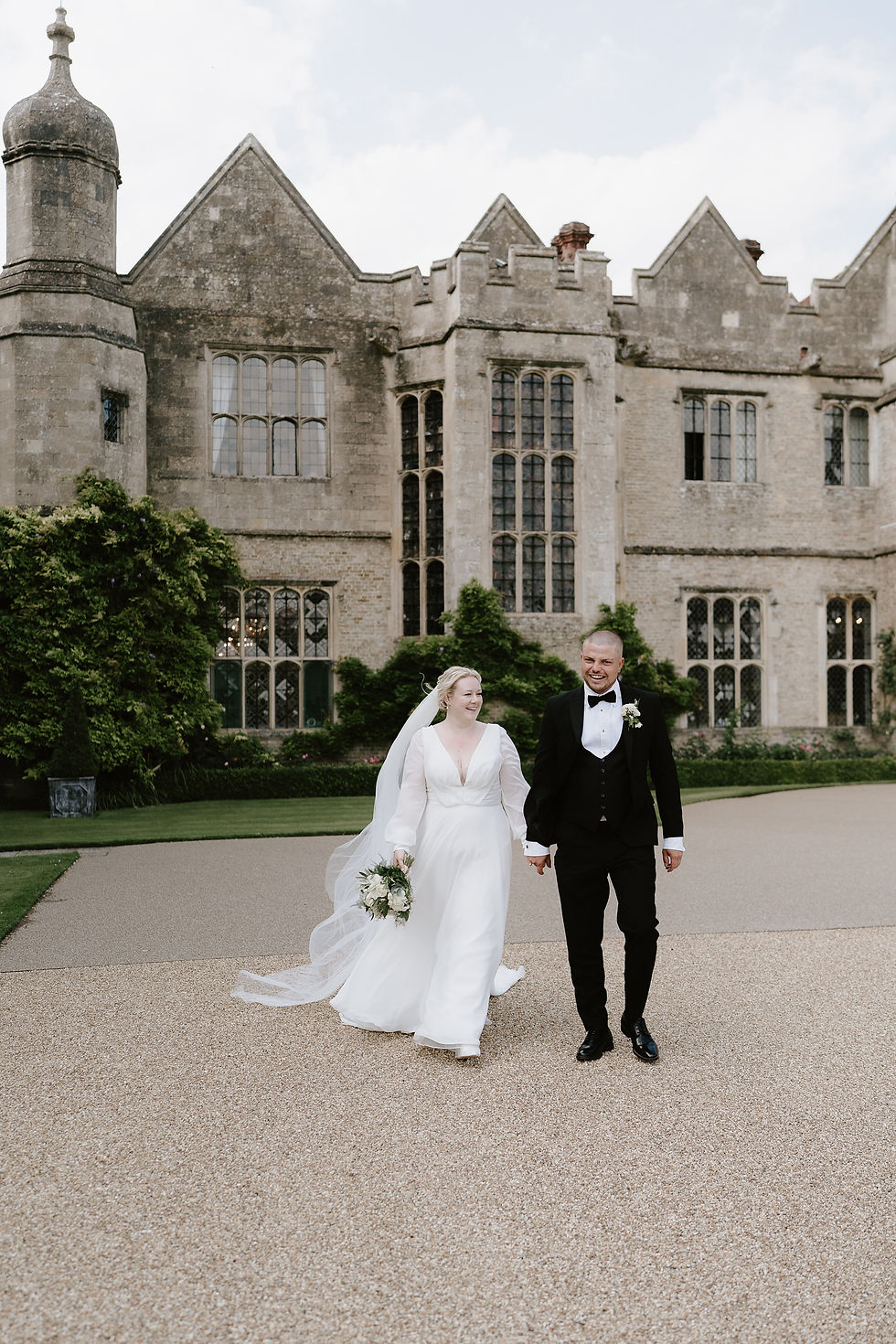 A bride and groom smile and hold hands as they walk in front of their Tudor house venue background