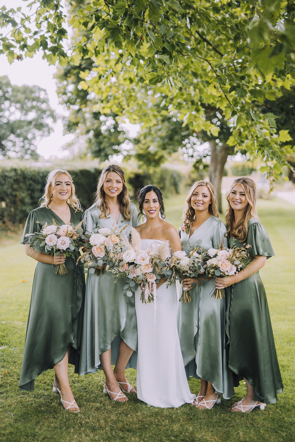The bride poses and smiles with 4 bridesmaids wearing green silk dresses and all holding bouquets