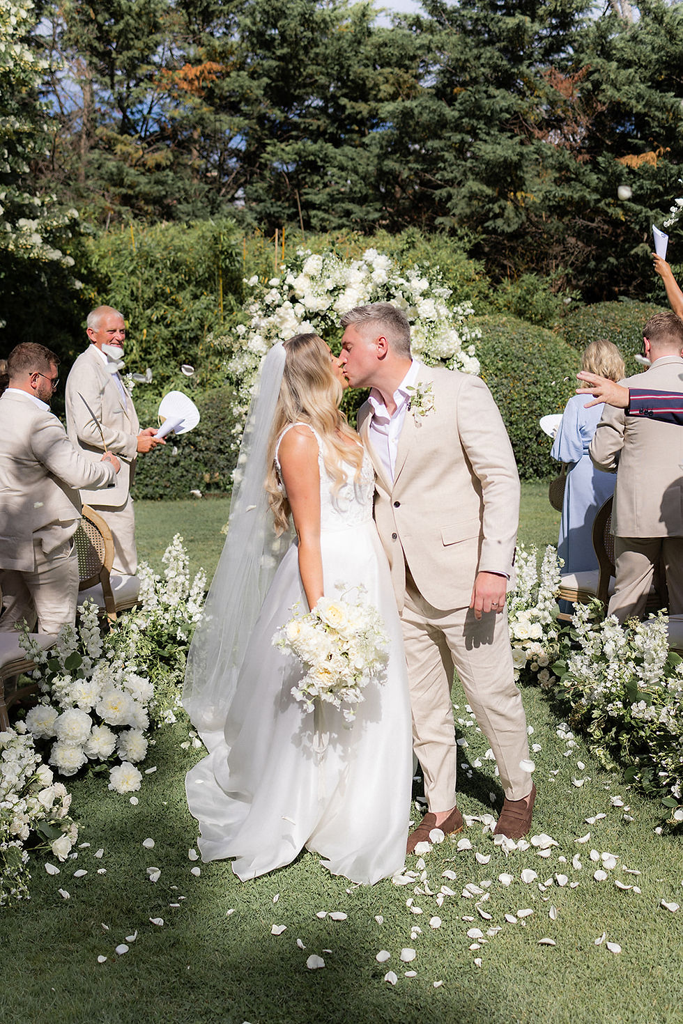 The bride and groom kiss half way down the ailse where guests have thrown confetti, with a floral arch backdrop