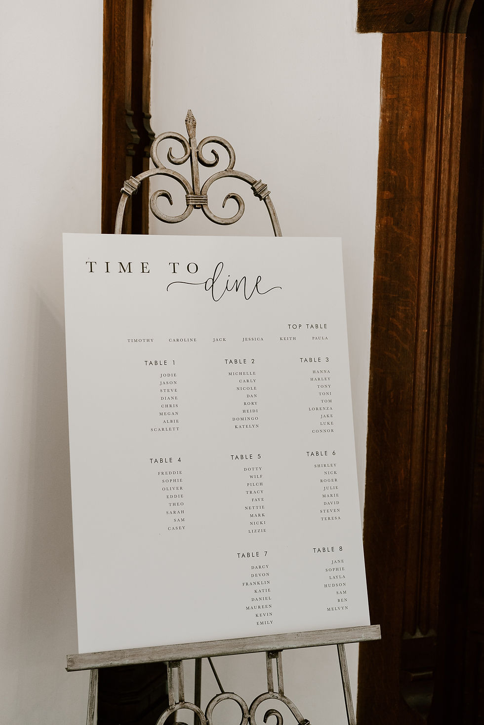 A wedding table plan sign with black calligraphy and text on white card, placed on a vintage style easel