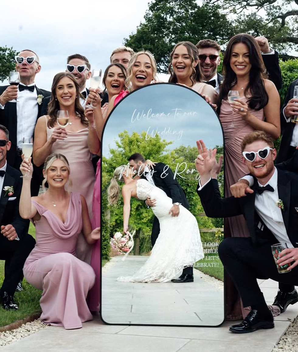 A bride and groom in the reflection of a wedding mirror sign, with the wedding party surrounding the mirror