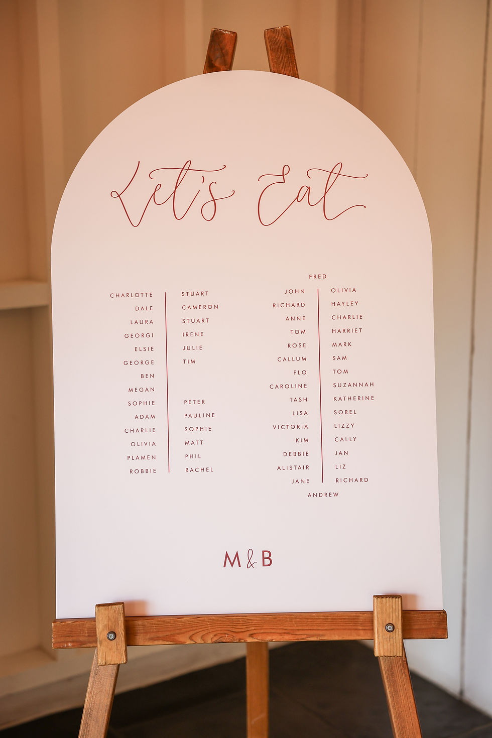 A wedding table plan with a calligraphy header saying 'Let's Eat' in burgundy on pale pink curved card. Placed on an easel.