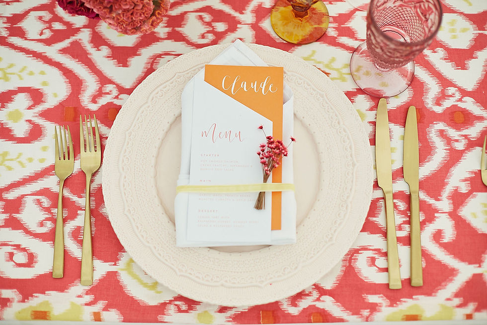 A white wedding menu with angled edges and orange calligraphy, layered on an orange place name personalised with white calligraphy. Placed on a pale pink plate which is on a bold coral and white tablecloth