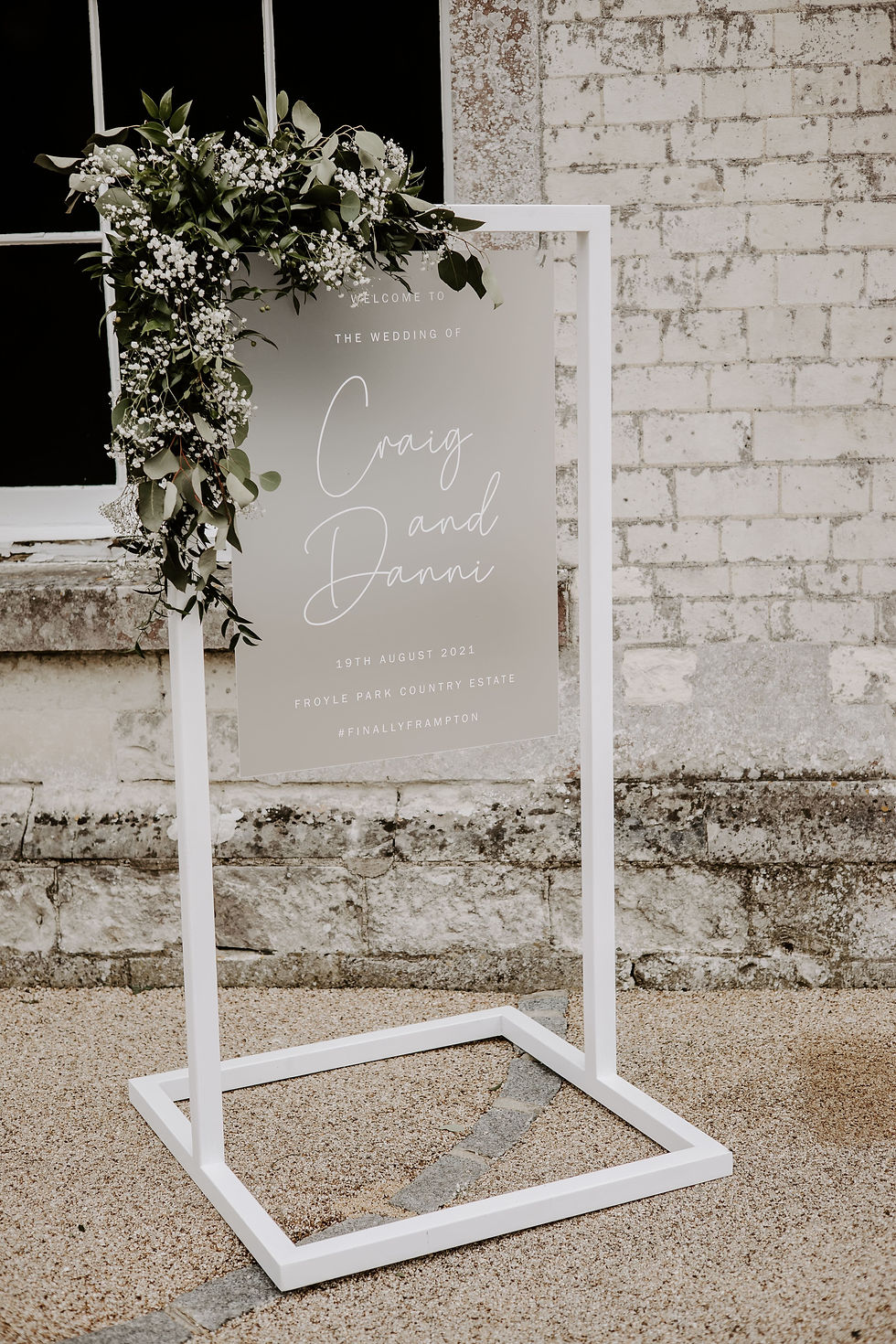 Wedding welcome plan with white calligraphy on a frosted Perspex background, hung on a white frame and styled with flowers.