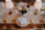 A wedding breakfast table styled with vintage lamps, a neutral tablerunner, gold cutlery, gold rimmed glasses, cream plates with pink edged detail and a romantic floral arrangement in pink, red and cream.