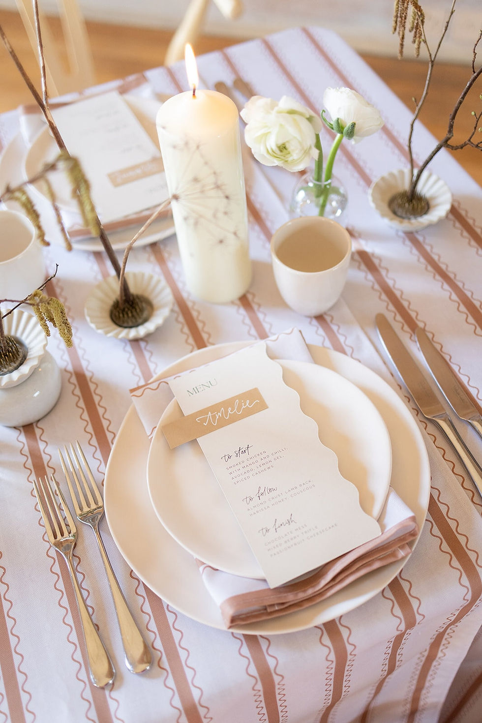 A wedding guest place name personalised with hand drawn calligraphy in white on a beige card, layered on a menu with waved edges and touches of calligraphy.